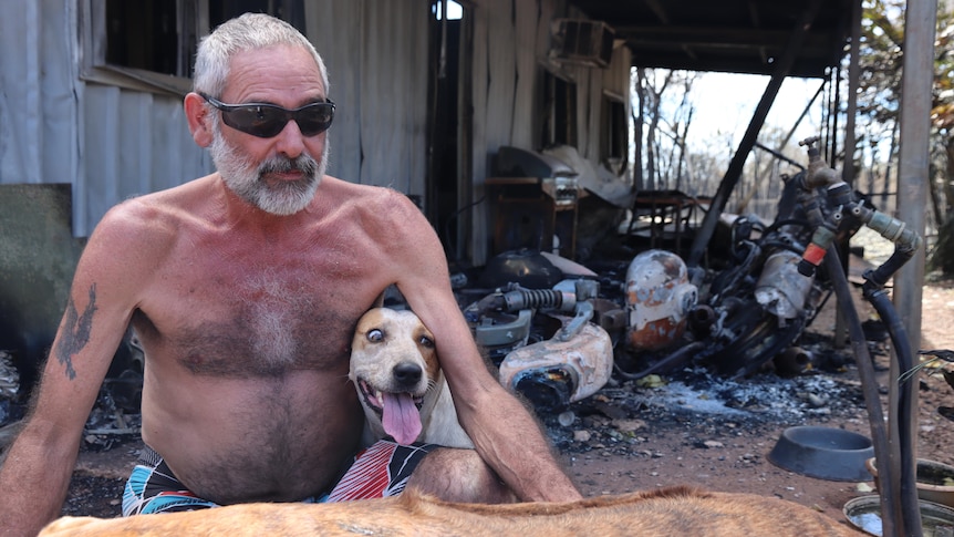 A shirtless man sits in front of a destroyed house with two dogs