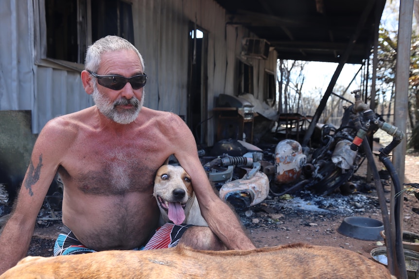 A shirtless man sits in front of a destroyed house with two dogs