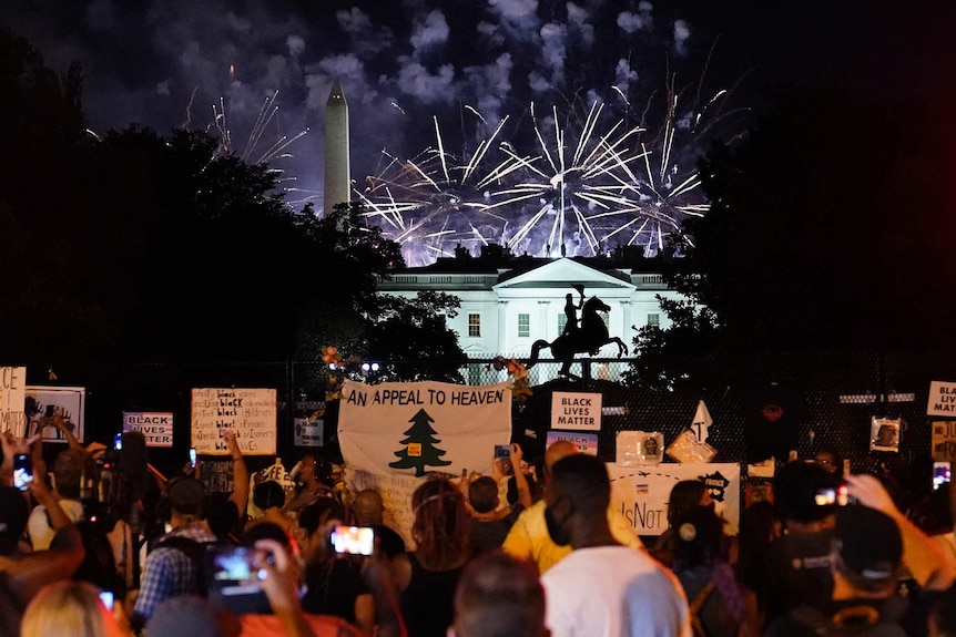 Bright lights colour the night sky above a white house with protesters carrying signs in front of it