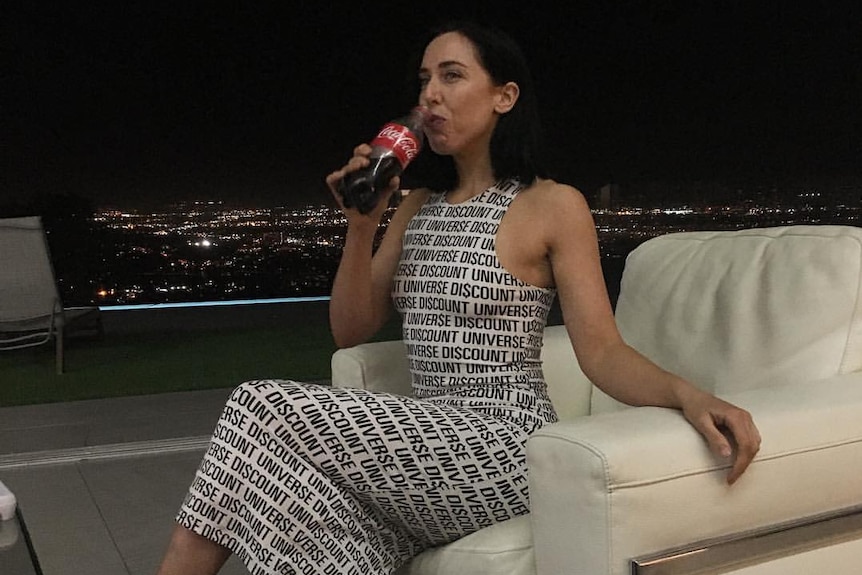 A woman in a dress printed with words sips a coke in a nighttime club on an armchair