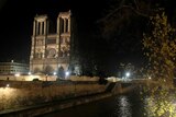 lights shine on the notre dame cathedral which is shown with a river in the foreground