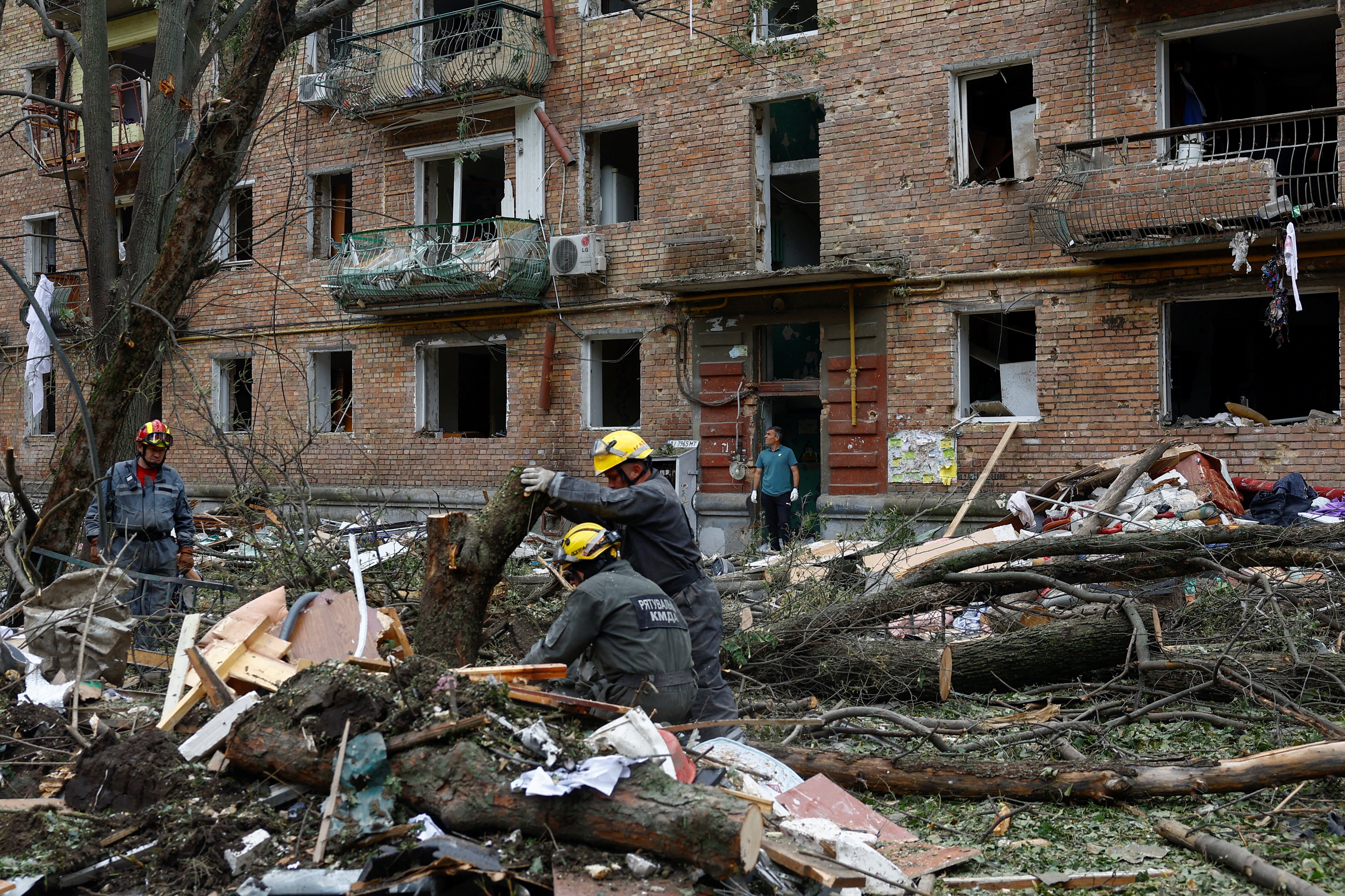 Ukrainian rescue workers in grey clothing and yellow hard hats sorting through rubble in front of a damaged brick building