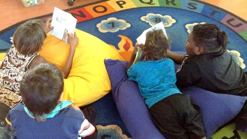 A group of children are lying down on a floor with cushions reading books together.