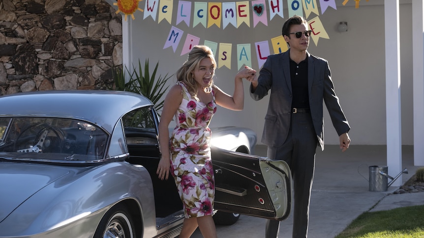 Blonde white woman in fitted floral dress holds hand of brunette white man in grey suit in front of welcome home sign.