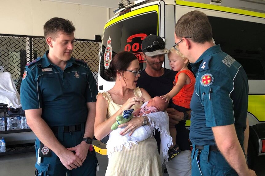 a man holding a child and a woman holding a baby speak with two ambulance officers