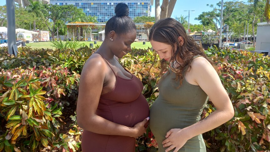 Two heavily pregnant girsl face each other laughing whilst their bellies touch each other