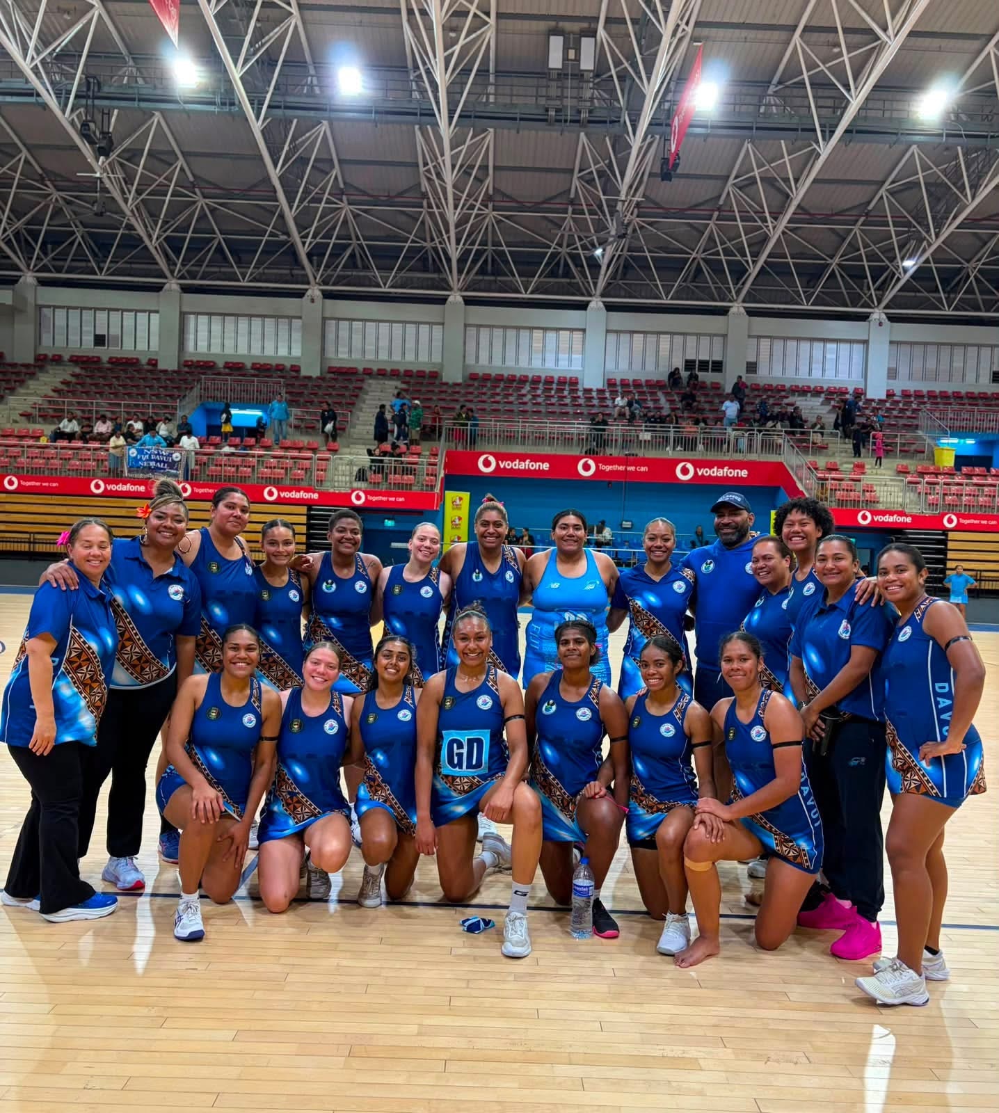 A group of young women and a few men wearing blue netball uniforms and track suits stand in a group on a netball court smiling 
