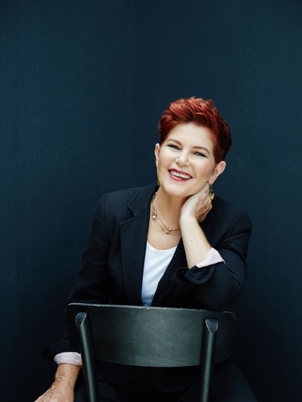 Portrait of a middle-aged woman with short red hair sitting on a seat smiling against a dark blue background.