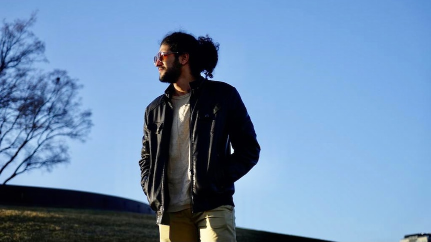 A man wearing a jacket, top and jeans with curly tied back long hair looks out in distance against blue sky