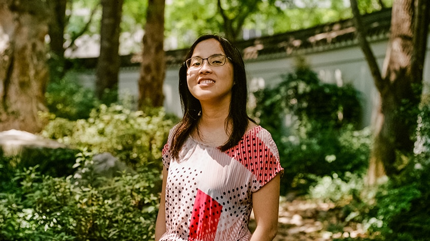 A young woman with glasses and in patterned dress stands smiling in lush green garden near white curved boundary wall.