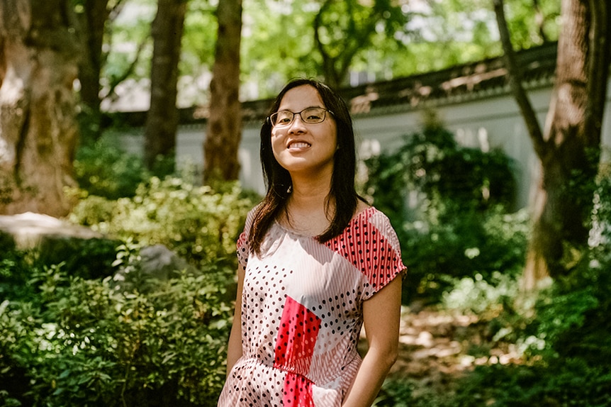 A young woman with glasses and in patterned dress stands smiling in lush green garden near white curved boundary wall.