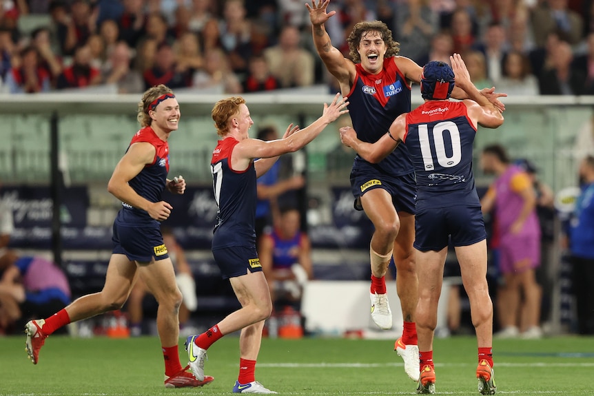 An AFL player leaps in the air with an arm raised in celebration after a goal, as his teammates gather.