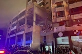 Orang-orang berkumpul di sekitar gedung bertingkat. Jendela-jendelanya telah pecah. Ini malam hari.