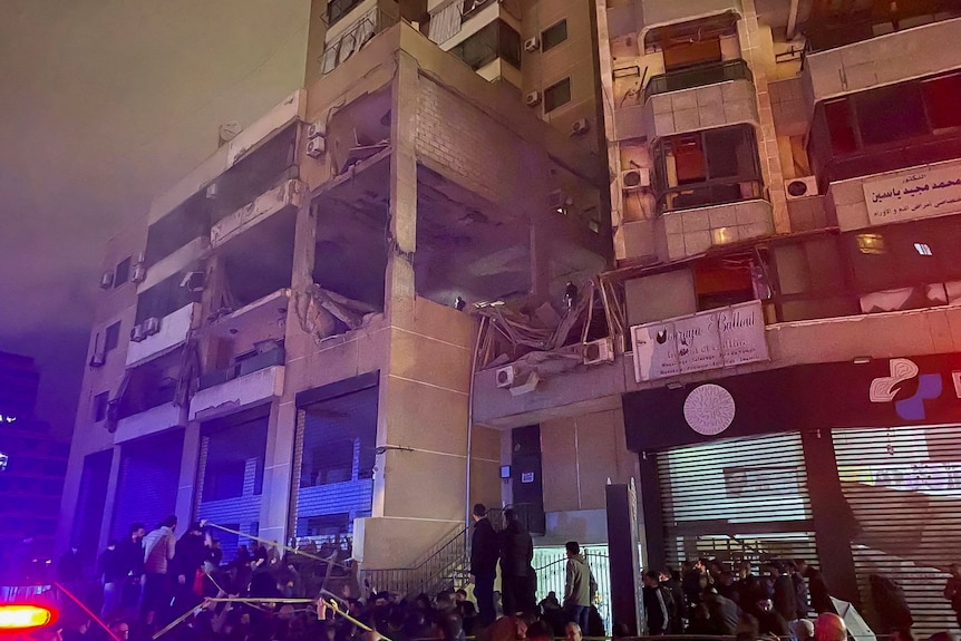 People are gathered around a multi-storey building. The windows have been blown out of it. It's nighttime.