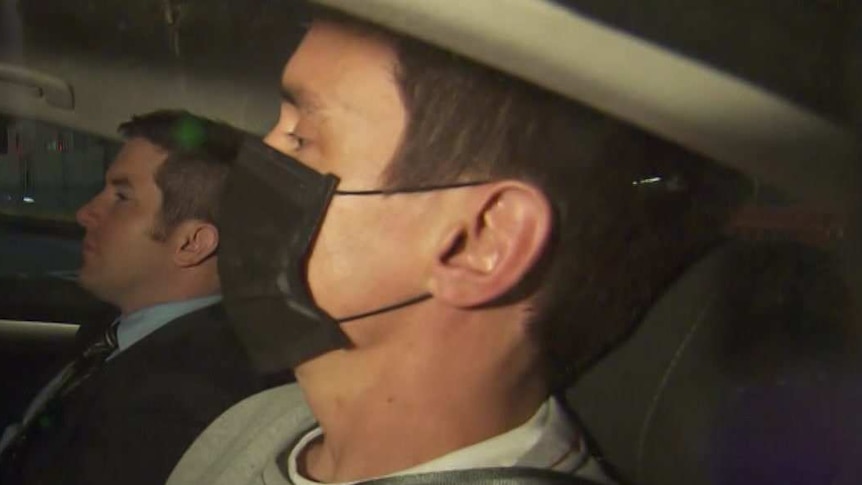 Richard Pusey, wearing a mask and grey jumper, is seated in the back passenger seat of a police car.