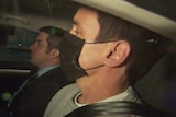 Richard Pusey, wearing a mask and grey jumper, is seated in the back passenger seat of a police car.
