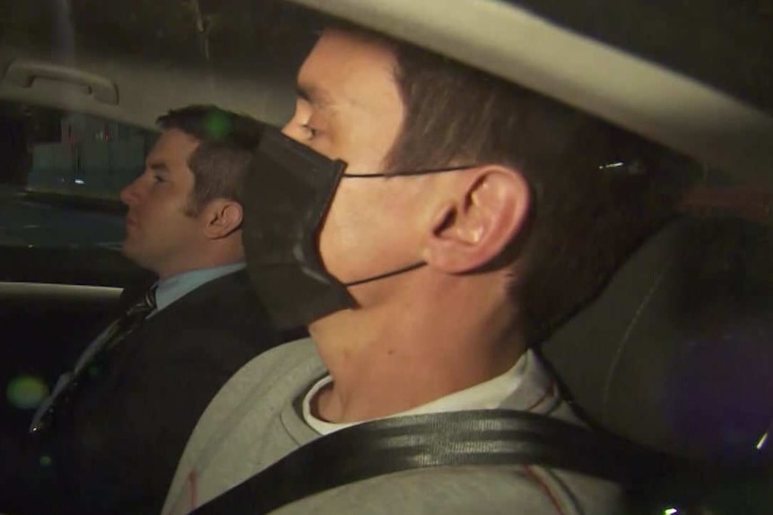 Richard Pusey, wearing a mask and grey jumper, is seated in the back passenger seat of a police car.