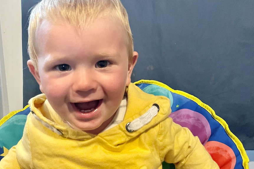 A blond toddler wearing a yellow hoodie smiling.