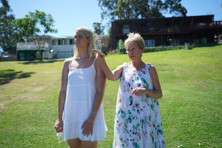 Two women in white dresses stand on a grassy hill.