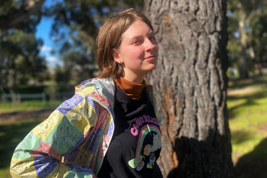 A teenager in a colourful jacket stands in front of a tree.