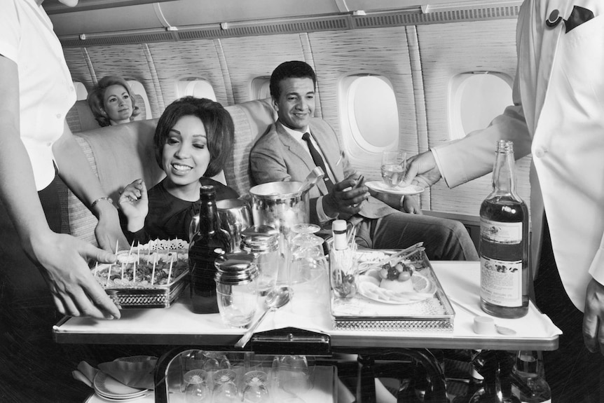 Flight crew serving food and beverages to passengers aboard an airplane, 1960s.