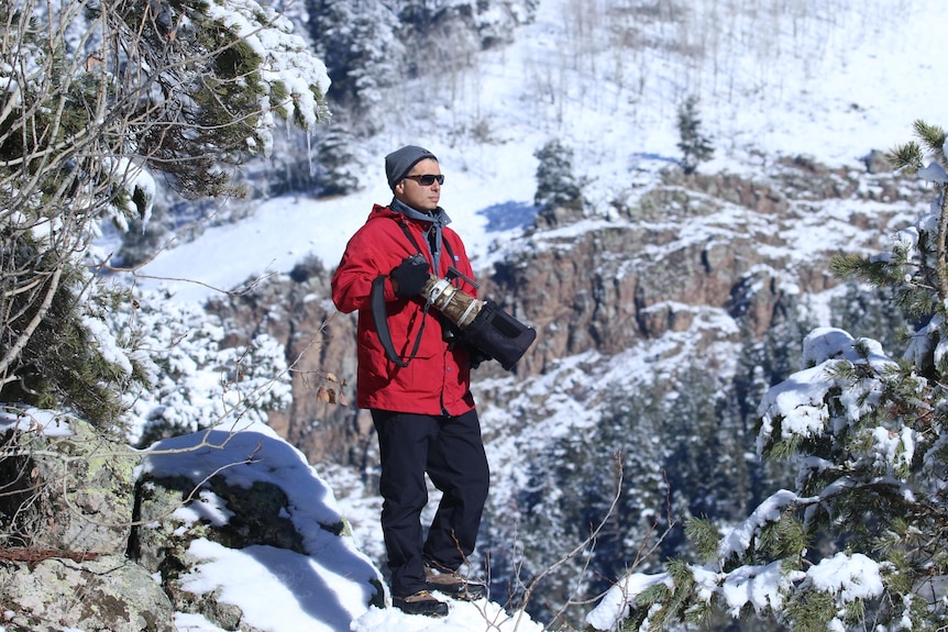 A man holds a large camera while walking on a snow covered mountian.