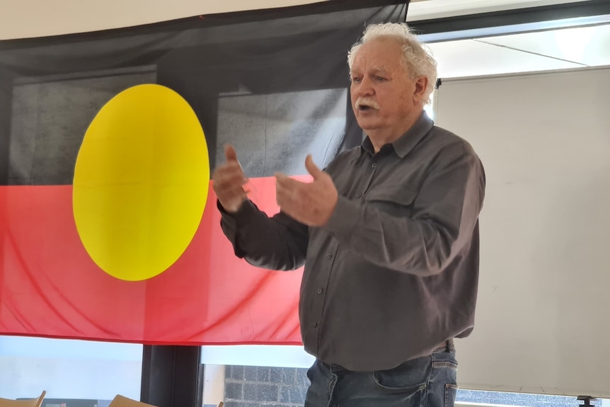A man stands in a room, speaking, behind him is the Aboriginal flag.
