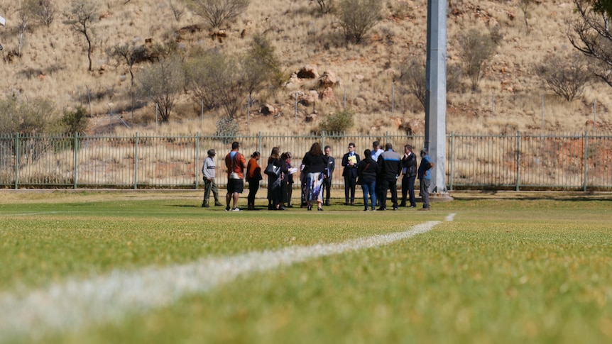 A low angle photo of a grass oval, with a group of people stood on the grass in the middle of the photo.