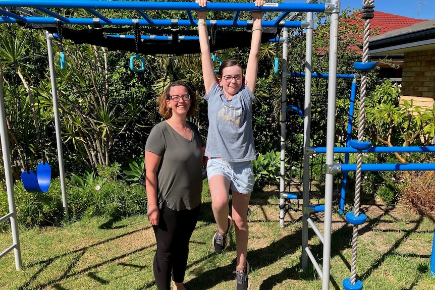 Thalia swings from a monkey bar, while her mum Diane stands next to her.