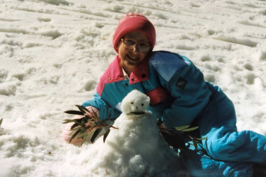 A girl in the snow, wearing cold-weather clothing, smiles as she sits behind a small snowman.