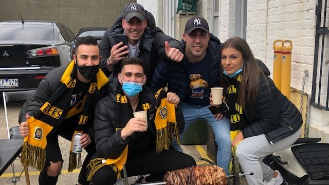 A group of five Richmond Tigers fans wearing black and yellow scarfs and posing around a slab of kebab meat over some coals.