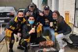 A group of five Richmond Tigers fans wearing black and yellow scarfs and posing around a slab of kebab meat over some coals.