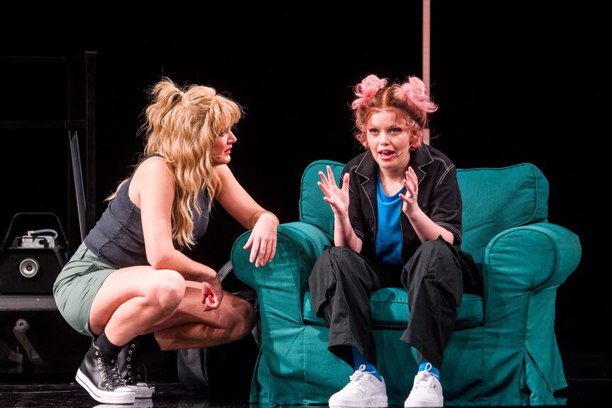 Two actors on stage, one young woman crouches next to another young woman with pink hair sitting in a green armchair