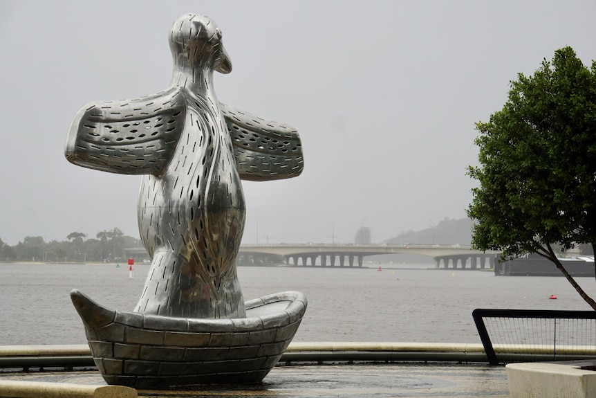 A silver statue of a bird next to a wide river with a traffic bridge in the background on a rainy day.