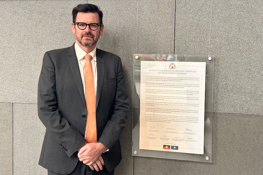 A middle-aged caucasian man in a grey suit and an orange tie stands next to a plaque on a wall.