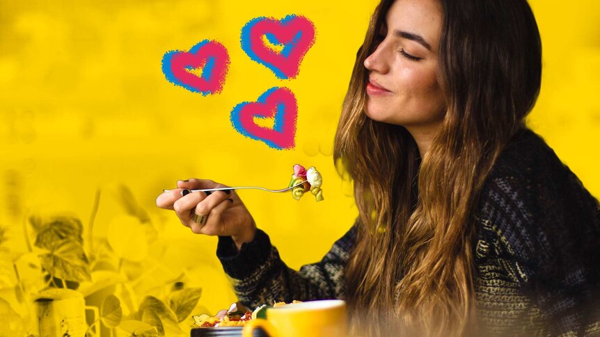 A woman sits alone eating a meal with her eyes closed and a smile on her face against a yellow background with hearts