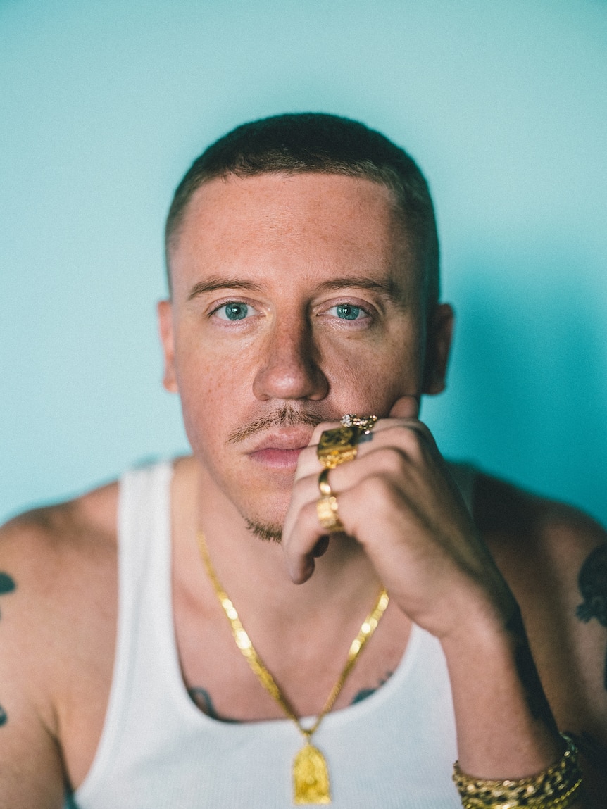 portrait of Macklemore wearing a singlet and gold jewelry with his left hand to his chin