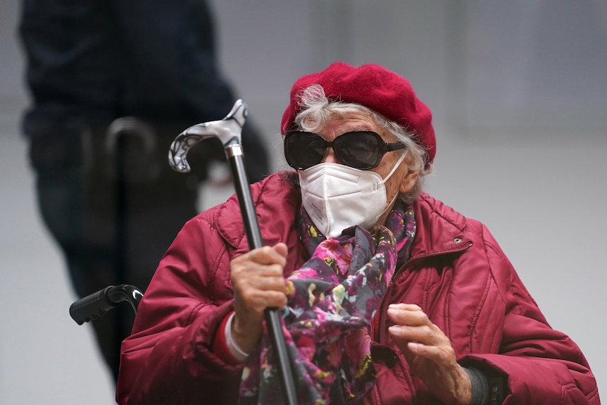 Irmgard F in sunglasses and masks sits in a wheelchair with a cane 