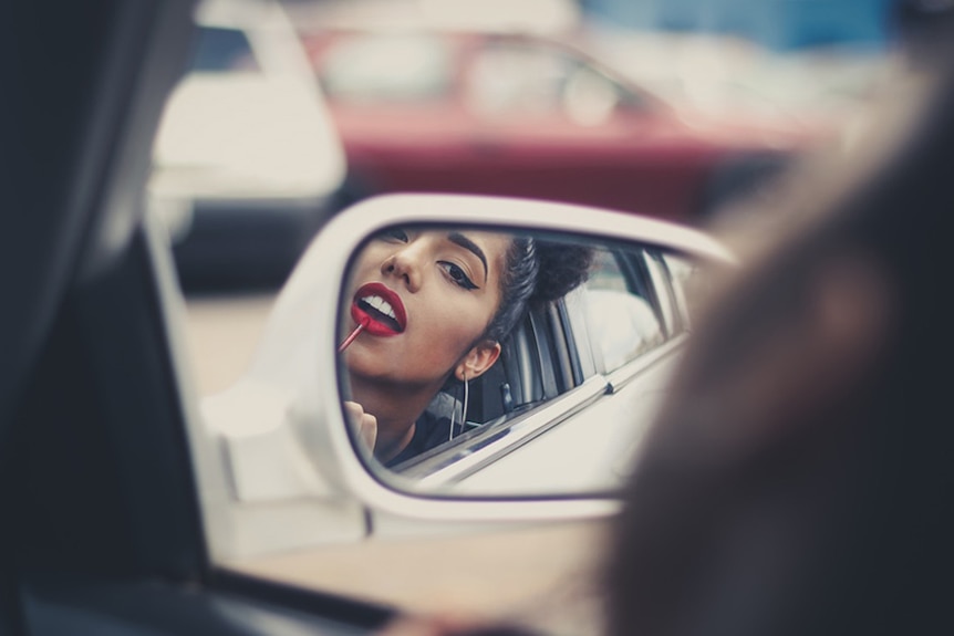 A woman puts on lipstick in her car mirror.