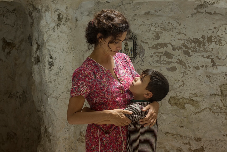 Penélope Cruz stands looking down at Asier Flores, both have arms wrapped around each other in a roughly painted room.