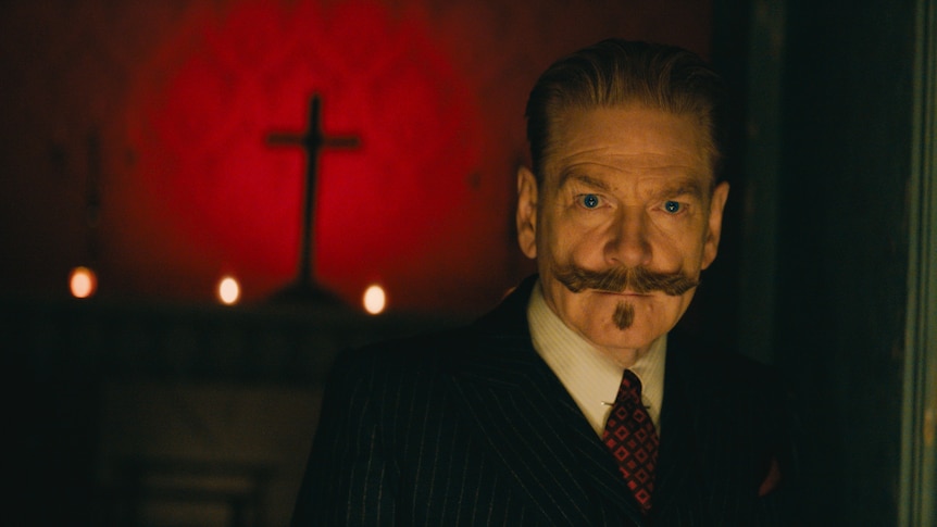 A older white man with a large moustache looks intensely, standing in front of a red background with a cross