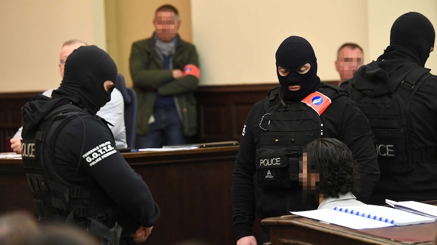 Guards with facemask stand next to Salah Abdeslam, whose face is pixelated.