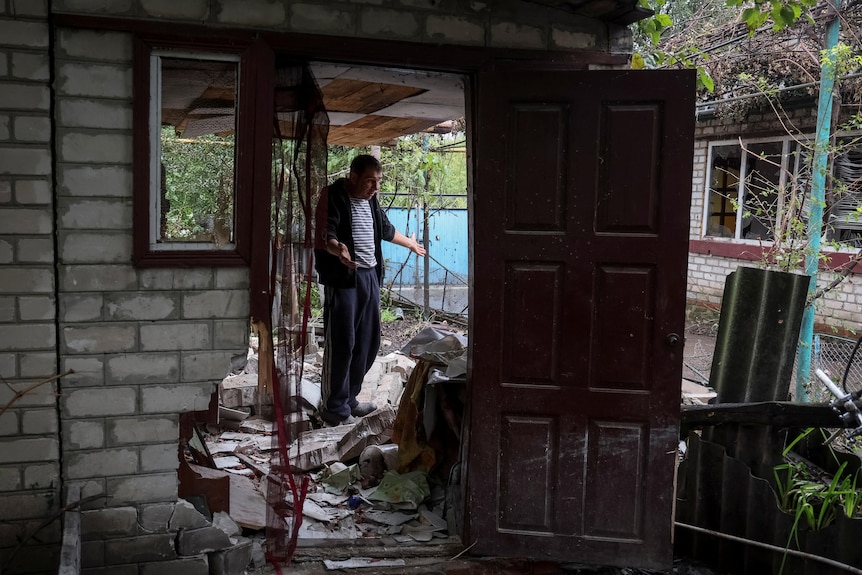 A middle-aged man in a striped shirt stands arms outstretched among the ruins of a grey brick house, its wooden door swung open.