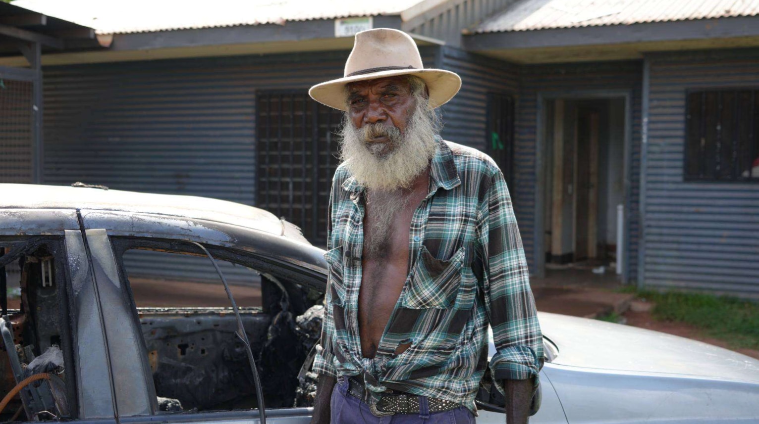 An older Indigenous man standing next to a burnt out car.