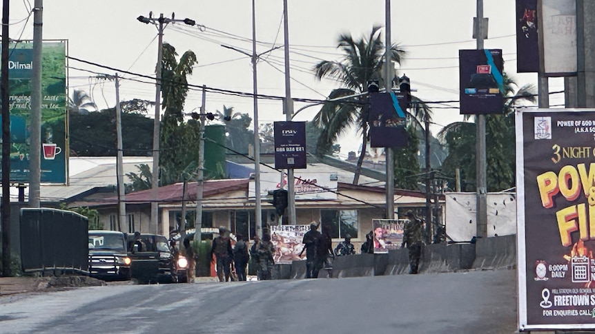 Men in military fatigues and holding guns stand in the distance on a road