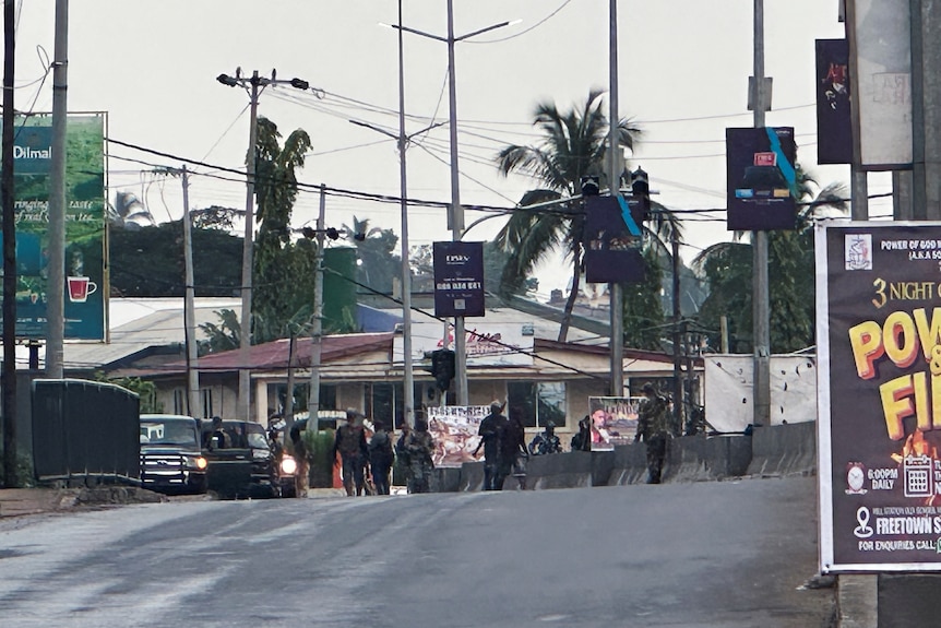 Men in military fatigues and holding guns stand in the distance on a road