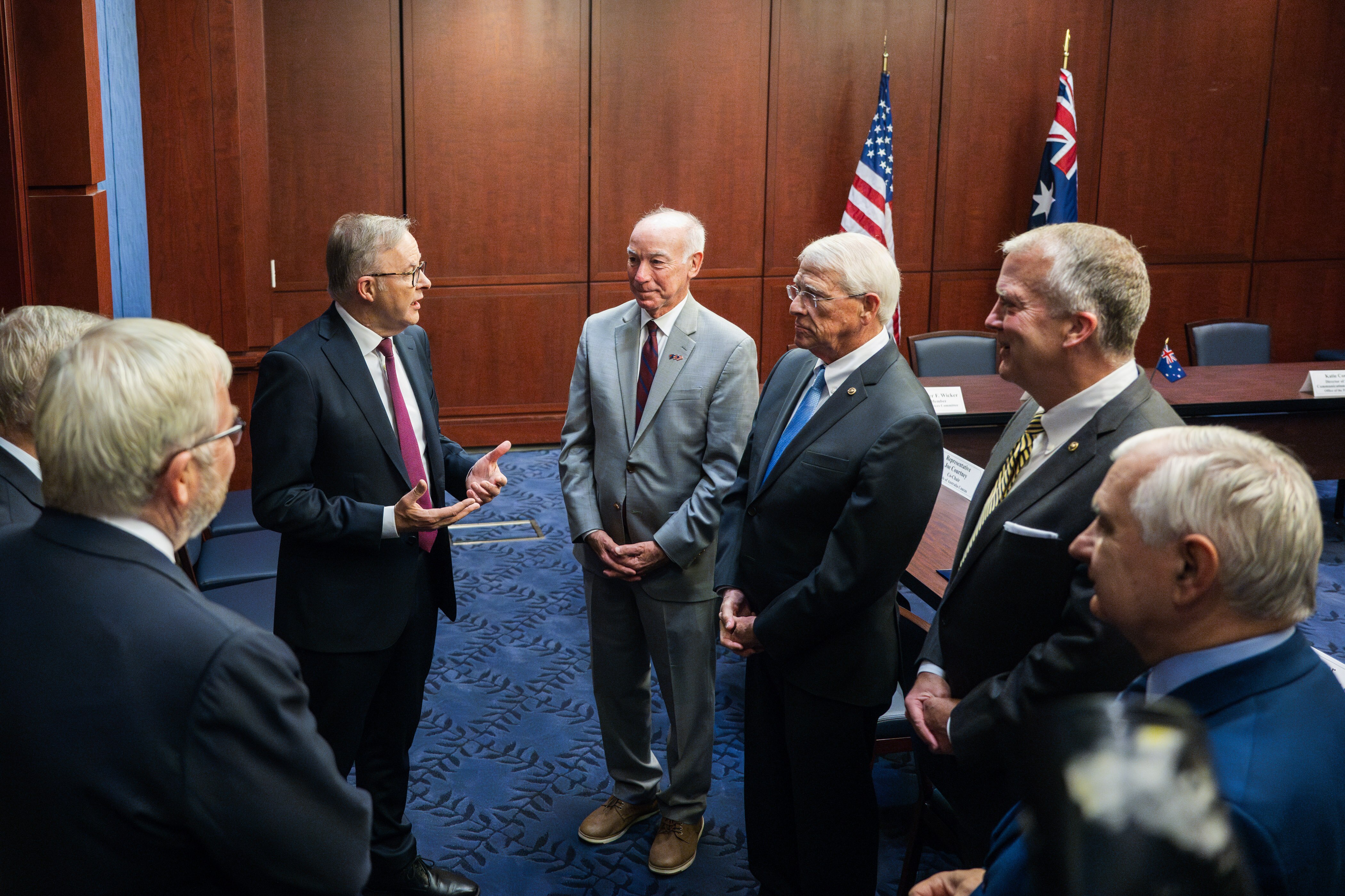 A group of men stands in a room, speaking. There are US and Australia flags next to a table behind them.