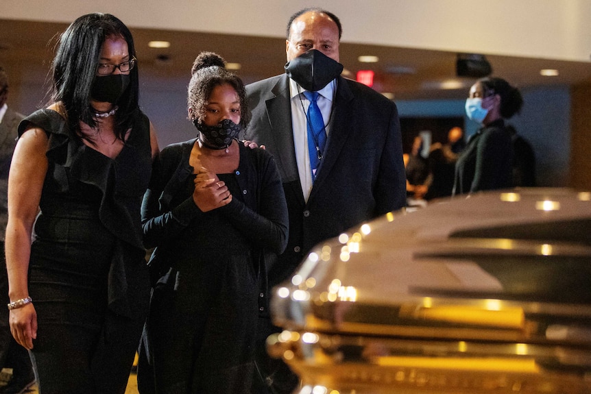 Two African American women and a man look at a gold coffin while wearing black masks