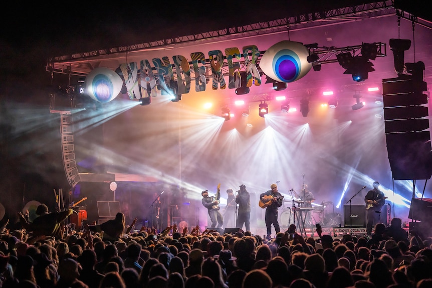 A bands plays on a stage at night lit up with pink lights in front of a big crowd.