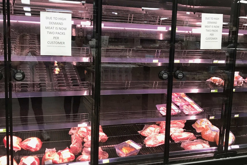 A supermarket fridge of meat is almost completely empty with a sign on the door limiting people to two packs each.